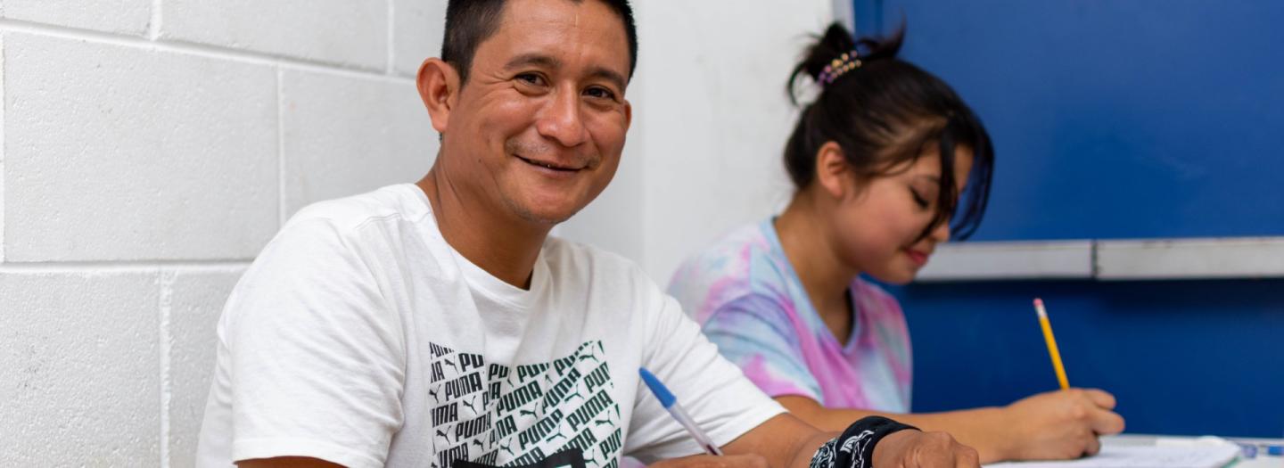 Ein Mann im T-Shirt sitzt an einem Pult und lächelt in die Kamera. In der Hand hat er einen Kuli, vor ihm liegt ein Heft. Im Hintergrund schreibt eine Frau in ein Heft.