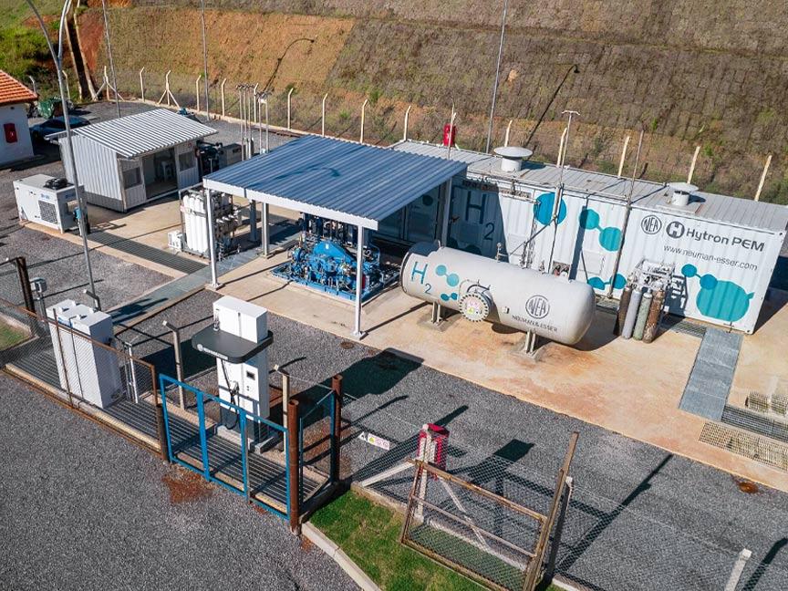 Eine eingezäunte Pilotanlage zur Produktion von grünem Wasserstoff steht am Rande eines Berghangs. Auf einem der verschiedenen, miteinander verbundenen Kessel ist die Aufschrift H2 zu lesen.