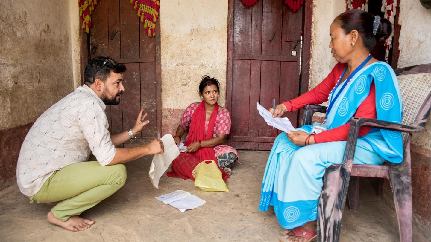 Drei Personen sitzen vor einer Hauswand auf dem Boden bzw. auf einem Stuhl und besprechen Papiere; eine Frau in traditioneller Kleidung mit blauem Sari schreibt Notizen, während die anderen zuhören und Dokumente in den Händen halten.