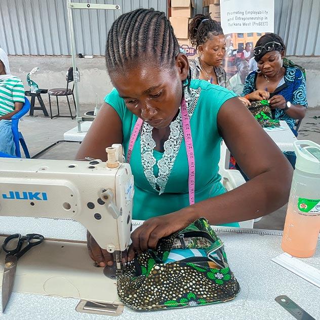 A woman works carefully at a sewing machine to make a bag from recycled materials; in the background are other women working on similar projects.
