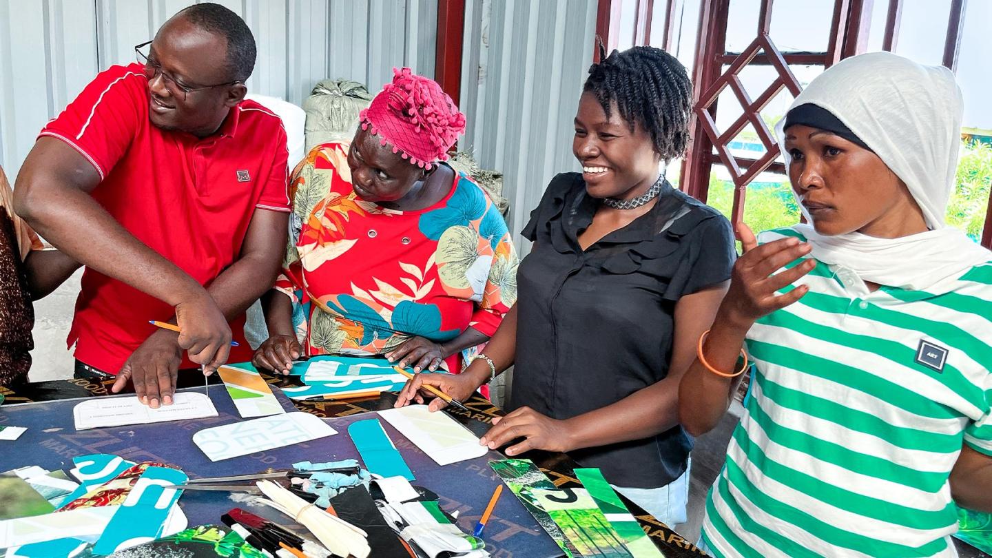Three women and one man stand together at a table; they are cutting and shaping materials that are laid out on a work surface.