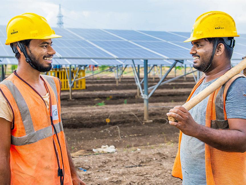 Zwei Männer mit Schutzhelmen und Warnwesten stehen lachend auf einem Solarfeld, einer hält eine Schaufel über der Schulter.
