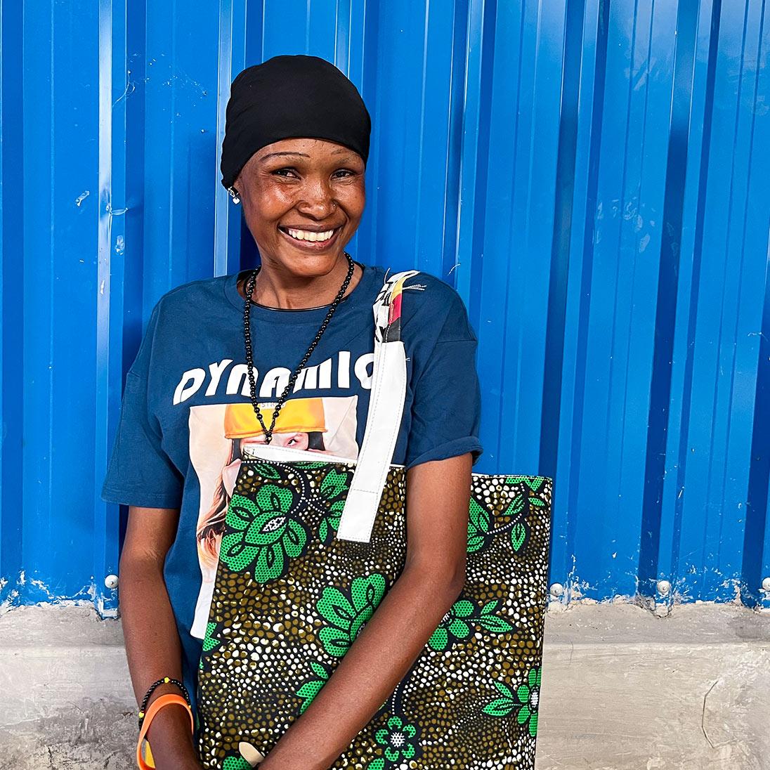A smiling woman stands in front of a blue metal wall wearing a printed t-shirt and carrying a brightly coloured fabric bag with a floral pattern on it.