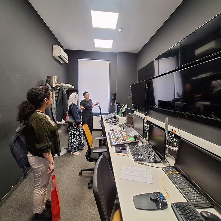 Three women are standing in a room with computers, where large screens hang on the walls.