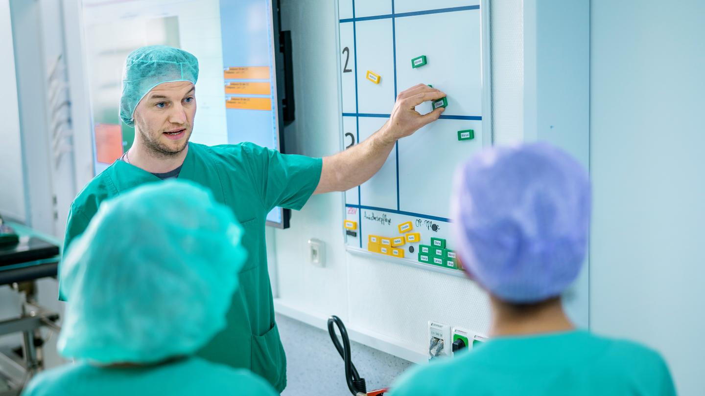 A man in surgical attire explains something to two team members in front of a planning board.