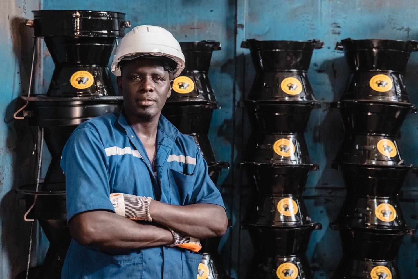A man in a white helmet stands in front of a wall where cookers are piled up.