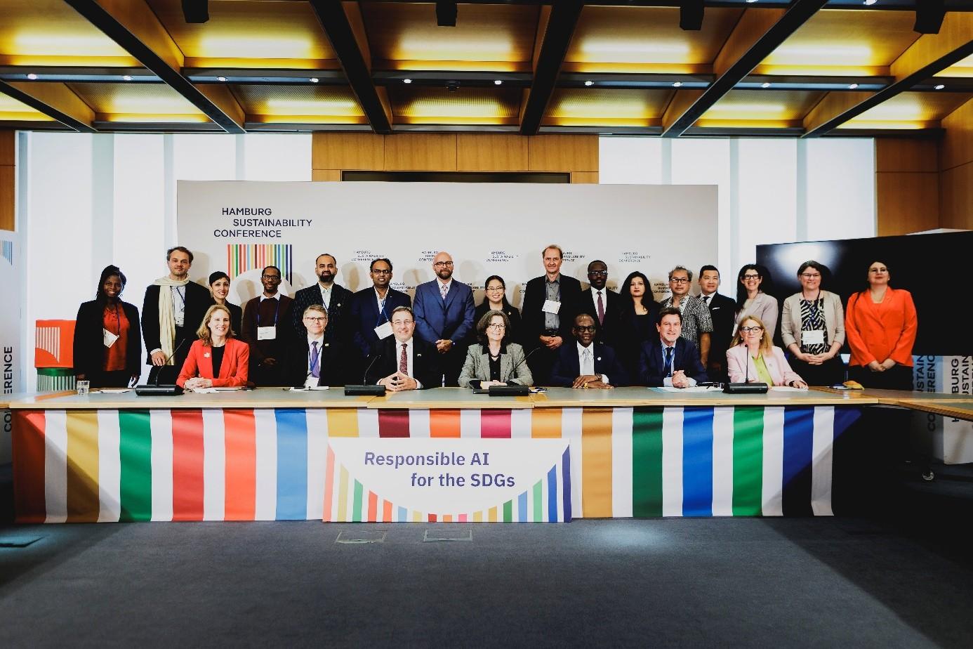 Eine Gruppe von Menschen steht in zwei Reihen vor einem langen Tisch, an dem ein bunt gestreifter Banner befestigt ist. 