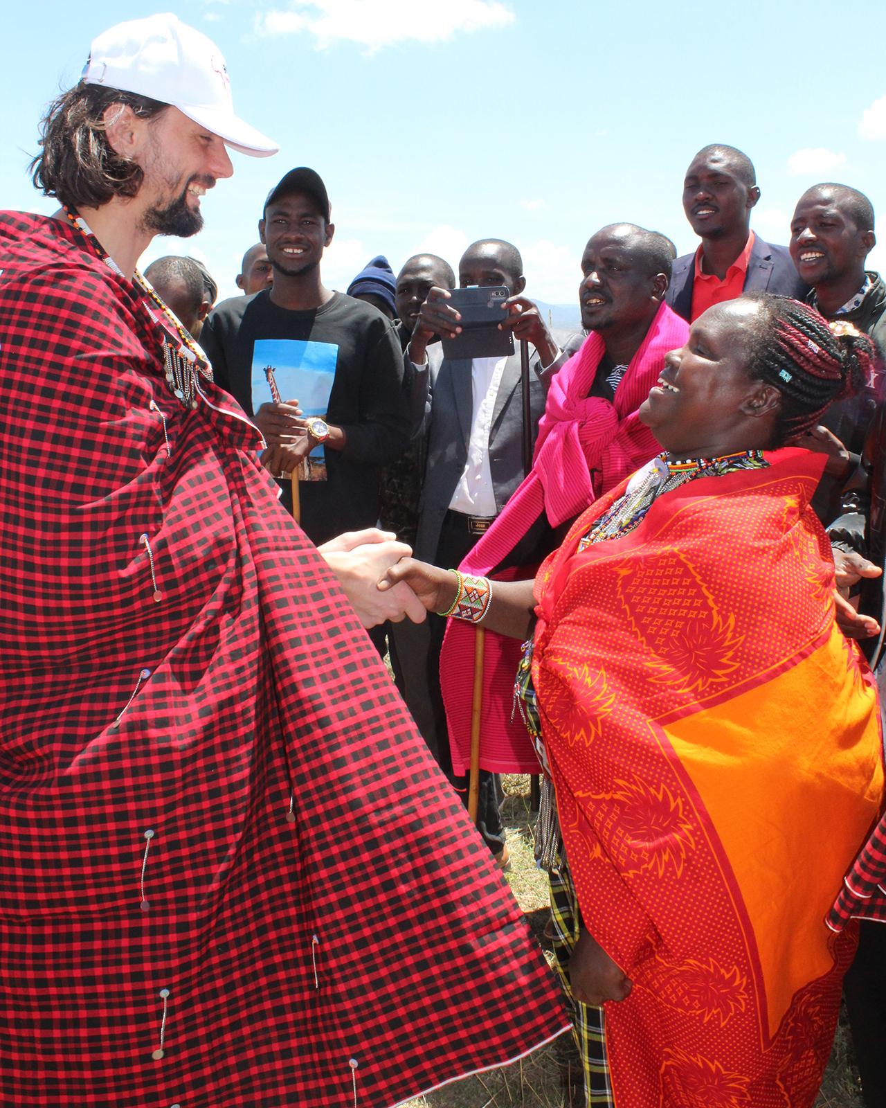 Ein Mann in traditioneller rot-schwarz karierter Kleidung und weißer Kappe begrüßt lächelnd eine Frau in einem orangefarbenen Gewand, umgeben von einer Gruppe von Menschen im Freien.