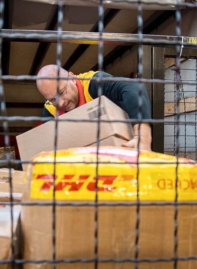 Ein Mann in Arbeitskleidung hebt in einem Lager zwischen Paketen einen Karton an, im Vordergrund ist ein Gitter unscharf zu sehen.