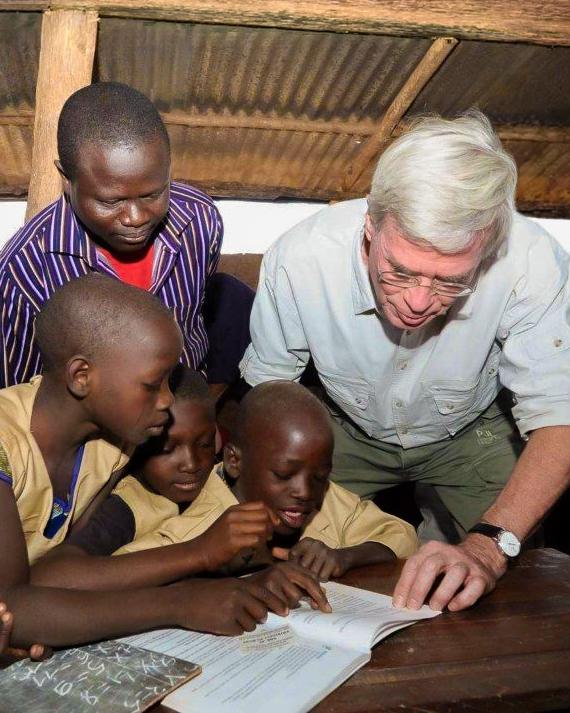 Ein Mann beugt sich über einen Tisch und zeigt mehreren Kindern in einer einfachen Hütte etwas in einem offenen Heft, während ein weiterer Erwachsener zuschaut.