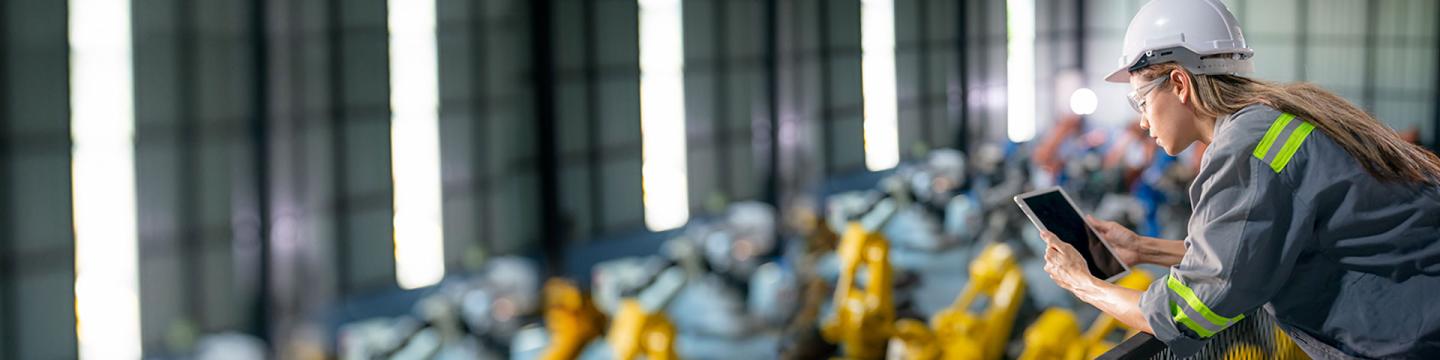 Eine junge Fabrikarbeiterin in Schutzkleidung schaut auf ein Tablet.
