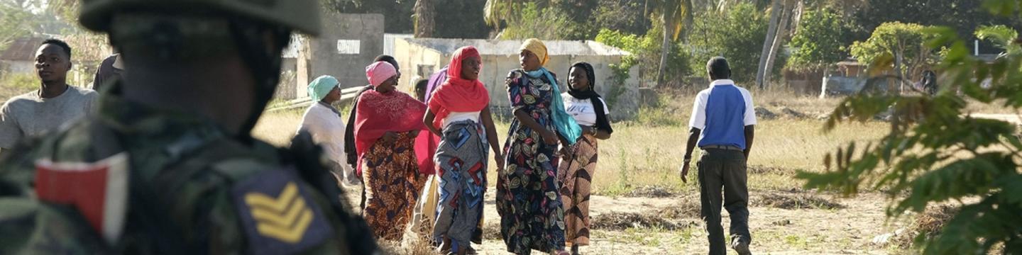 Mehrere Frauen laufen über einen Weg. Ein Soldat steht im Vordergrund und beobachtet die Szene.