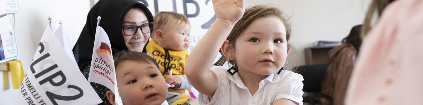 Eine Frau hält bei einer Veranstaltung ein Kleinkind, zwei weitere Kinder stehen im Vordergrund.