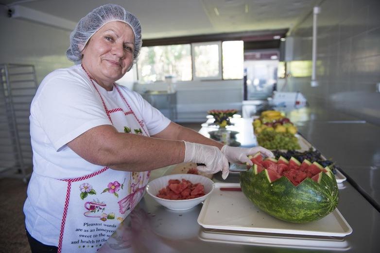 Eine Frau mit Haarnetz schneidet eine Wassermelone, im Hintergrund ist ein Buffet aufgebaut.