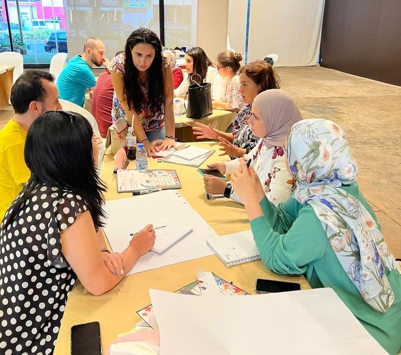 Flüchtlinge und aufnehmende Bürger*innen sitzen bei einem Arbeitstreffen zum Thema Konfliktlösungen an Tischen. Vor ihnen liegen Notizbücher und Arbeitsmaterialien.