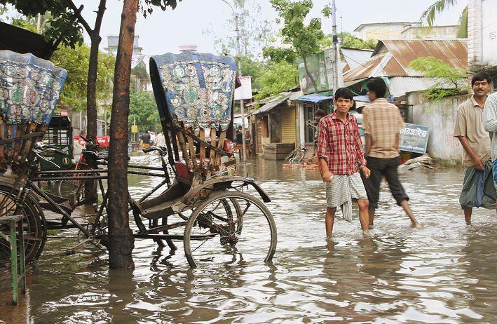 Vier Personen laufen durch kniehohes Wasser durch eine überschwemmte Straße in Bangladesh. Zwei Personen blicken in die Kamera.