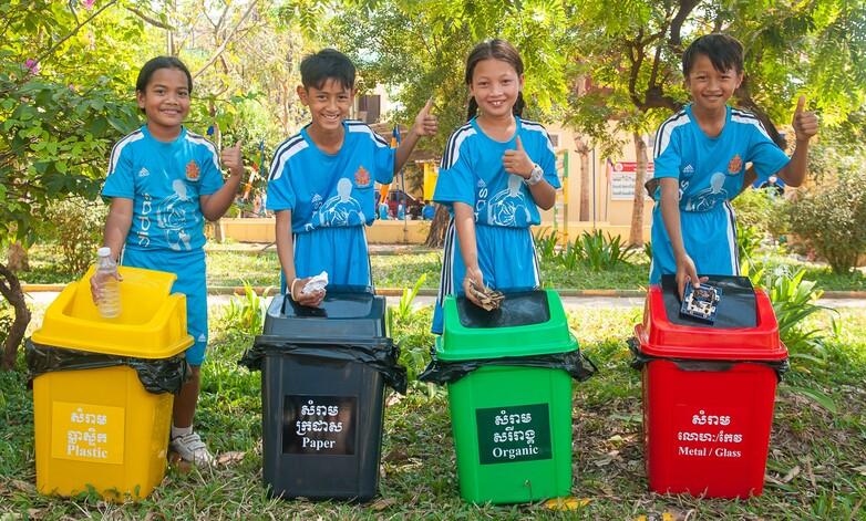 Vier Schüler*innen zeigen Mülleimer für Mülltrennung auf einem Pausenhof. Copyright: GIZ/Conor Wall