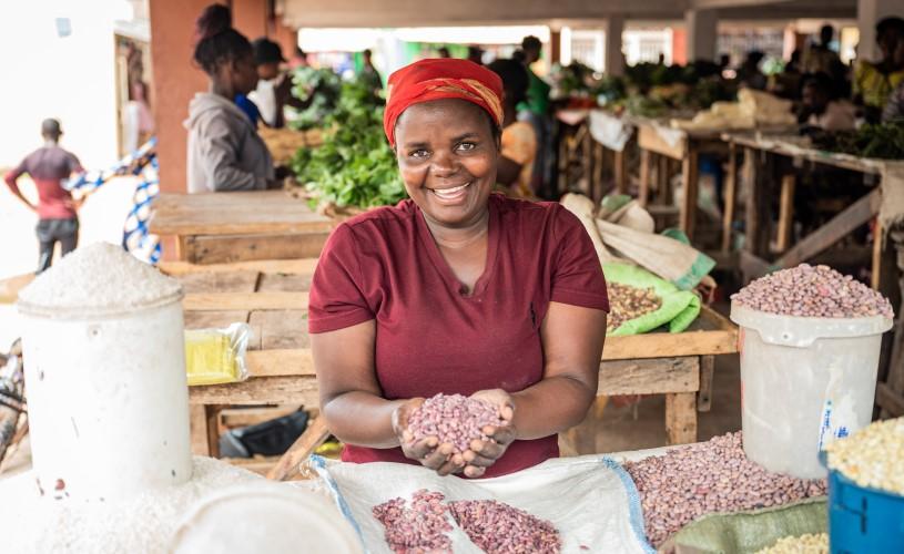 A woman smiles into the camera. Food is spread out on the floor in front of her to dry.