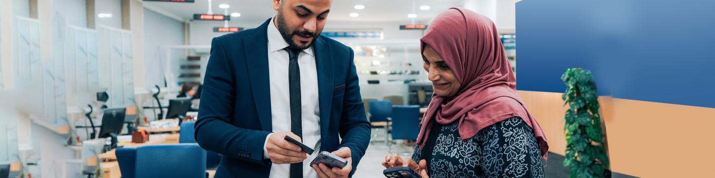 Ein Mann im Anzug hält in einer Bank ein Smartphone und eine Kreditkarte in den Händen und zeigt einer Frau, die ebenfalls ein Smartphone in der Hand hat, einen Vorgang.
