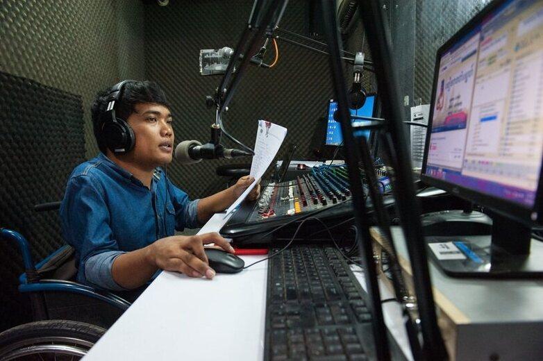A young man in a wheelchair is speaking into a microphone in front of the mixing desk at a recording studio.