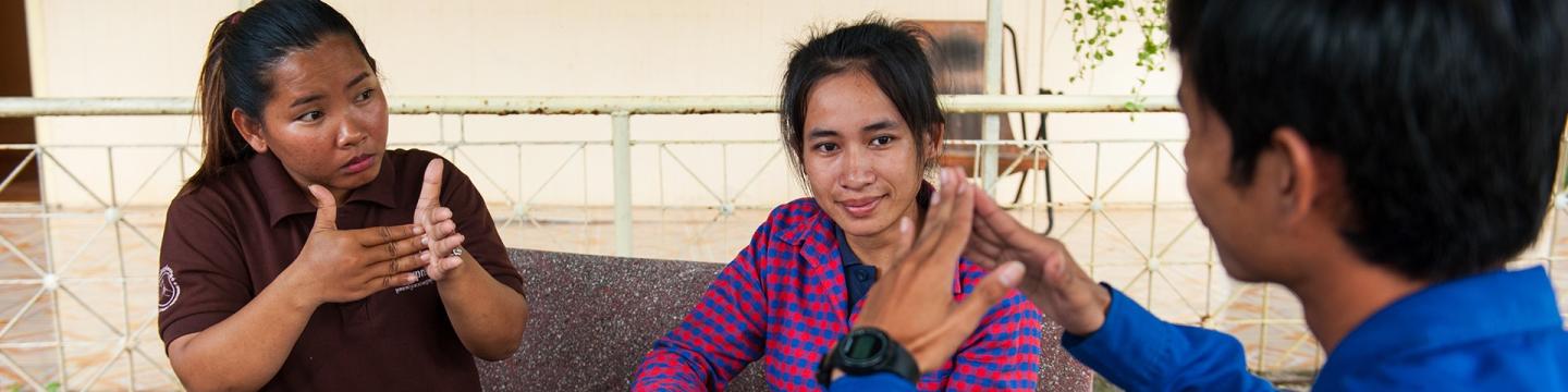 Zwei Personen kommunizieren in Gebärden. Zwischen ihnen sitzt eine dritte Person und lächelt in die Kamera.