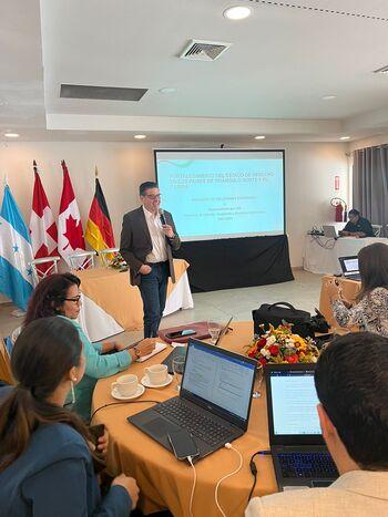 A man holding a talk at an event in front of an audience of people with laptops and notebooks.