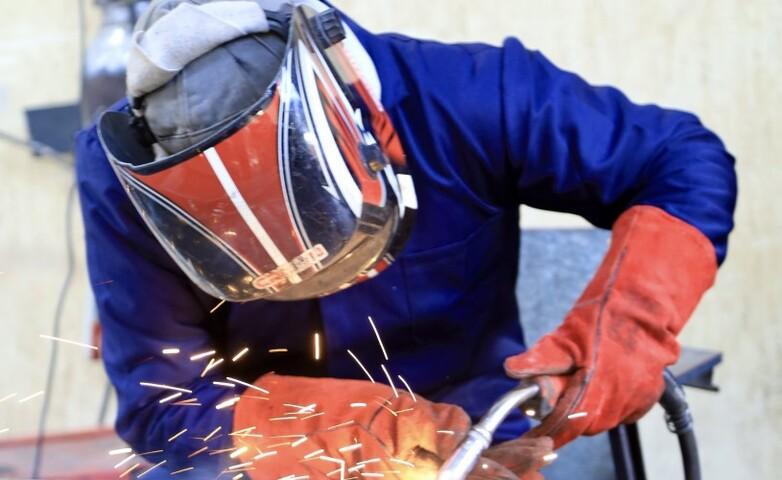 Eine Arbeitskraft in Schutzausrüstung mit Schweißhelm und Handschuhen schweißt in einer Werkstatt Metall, wobei helle Funken entstehen.