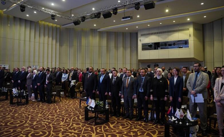 Attendees in formal attire standing in a conference room, with small flags on desks in the front row.