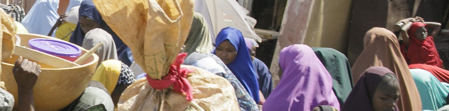 Dicht gedrängte Menschen auf einem Marktplatz. Einige tragen große Schüsseln und Waren.