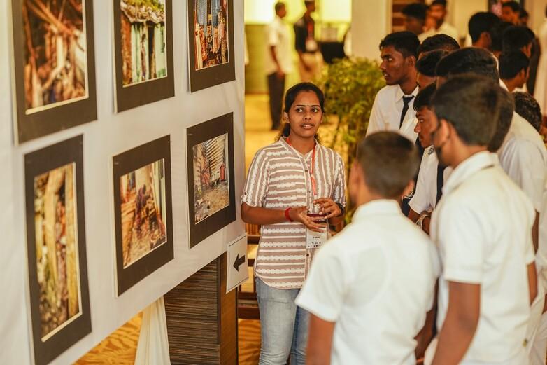 Eine junge Fotografin erklärt Schüler*innen, die vor einer Bilderwand stehen, ihre Fotografien.