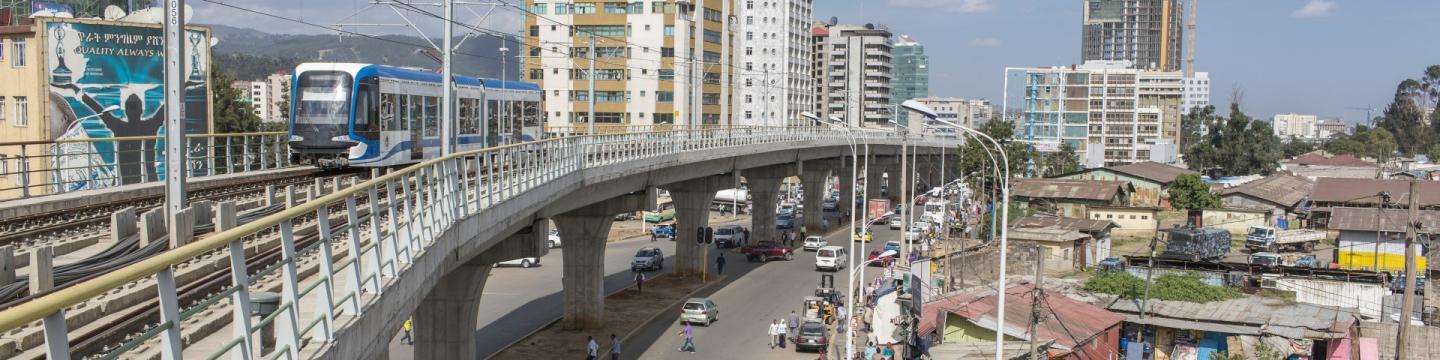 Ein blau-weißer Stadtbahnzug auf einer Hochbahnstrecke in einem belebten Stadtgebiet, umgeben von Hoch- und Flachbauten, bei klarem Himmel.