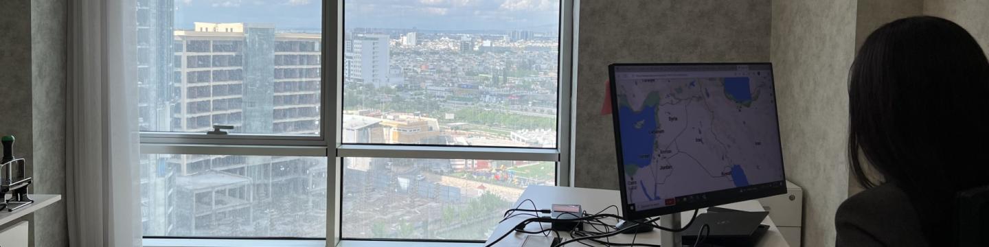 Eine Person arbeitet an einem Schreibtisch vor einem Monitor, auf dem eine Landkarte zu sehen ist. Ein großes Fenster bietet Ausblick auf eine Stadt.