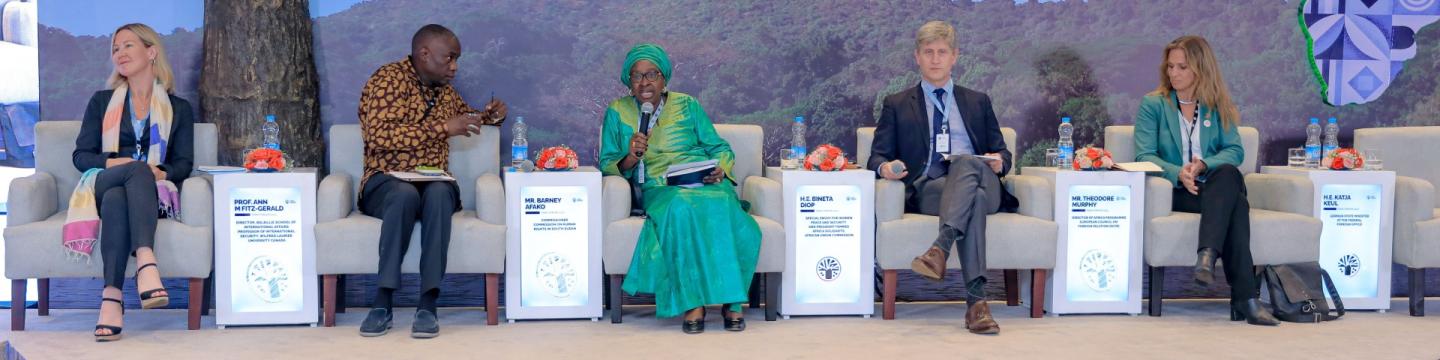 Eine Gruppe von Menschen sitzt auf einer Bühne, eine Frau hält ein Mikrofon.