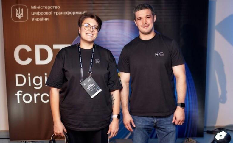 Two people stand in front of a banner of the Ministry of Digitalisation