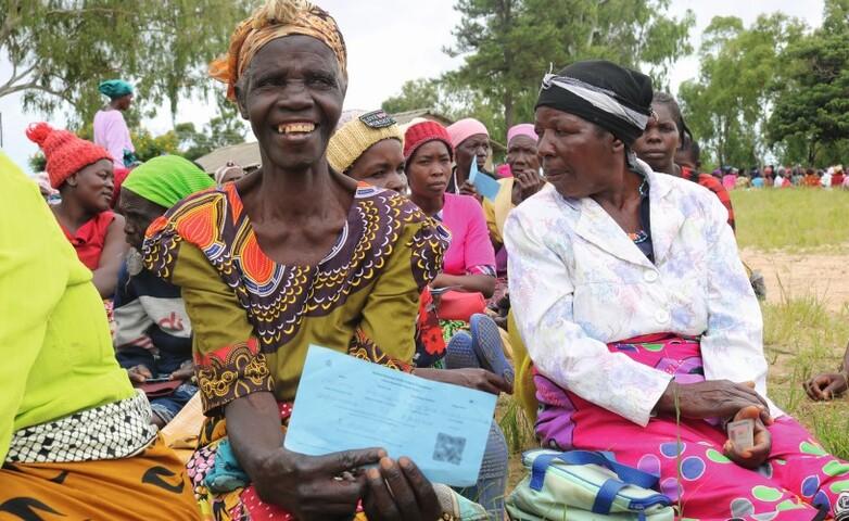 A woman holding her UBR card, sitting with other women in a meadow.