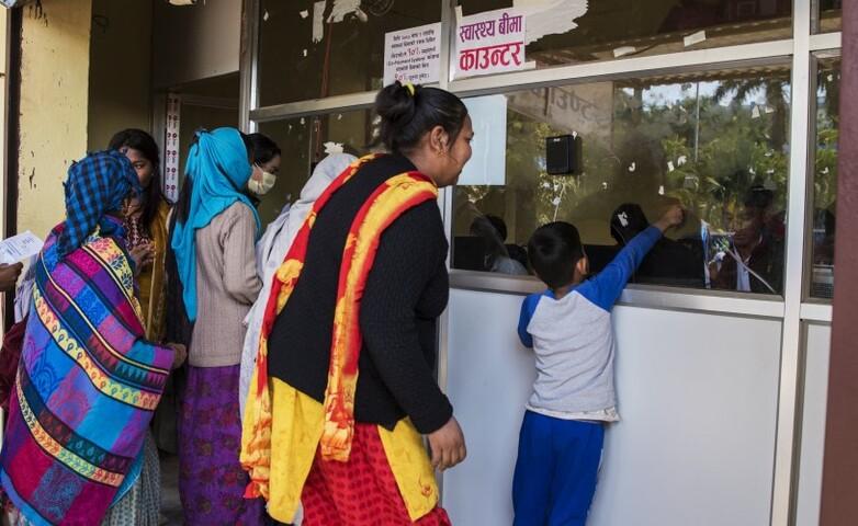 Menschen am Schalter einer Krankenkasse in Nepal, darunter Frauen und ein Kind, das in Richtung des Schalterfensters zeigt.