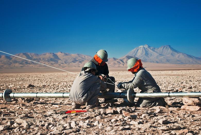 Tres personas vestidas con mono de trabajo efectúan trabajos en una tubería en un desierto rocoso.