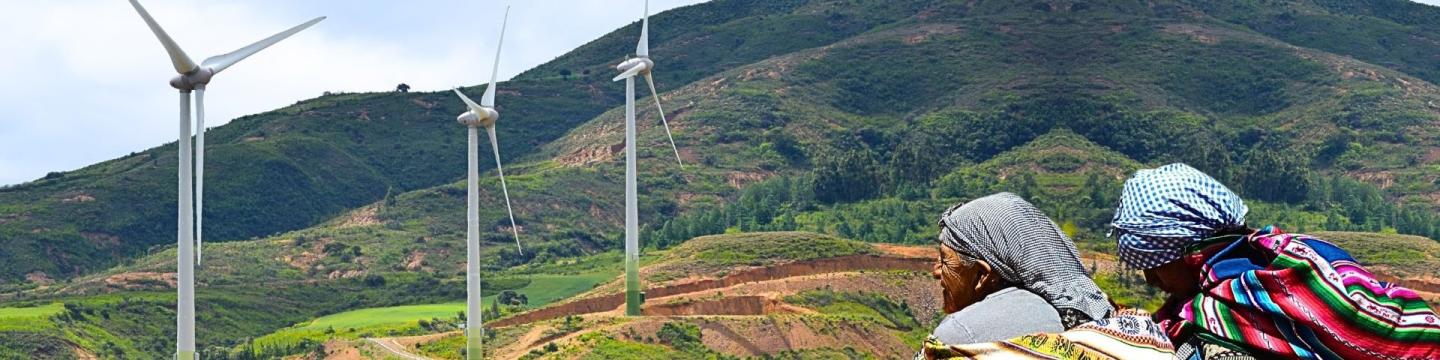 Dos mujeres bolivianas observan tres aerogeneradores en medio de un paisaje verde