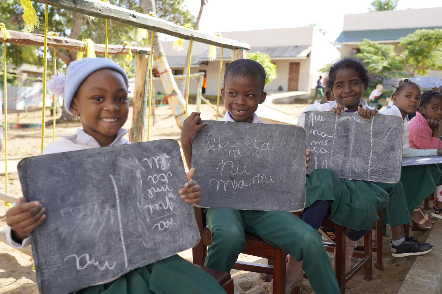 Grundschulkinder sitzen im Freien und zeigen Tafeln, auf denen sie lesen und schreiben üben.