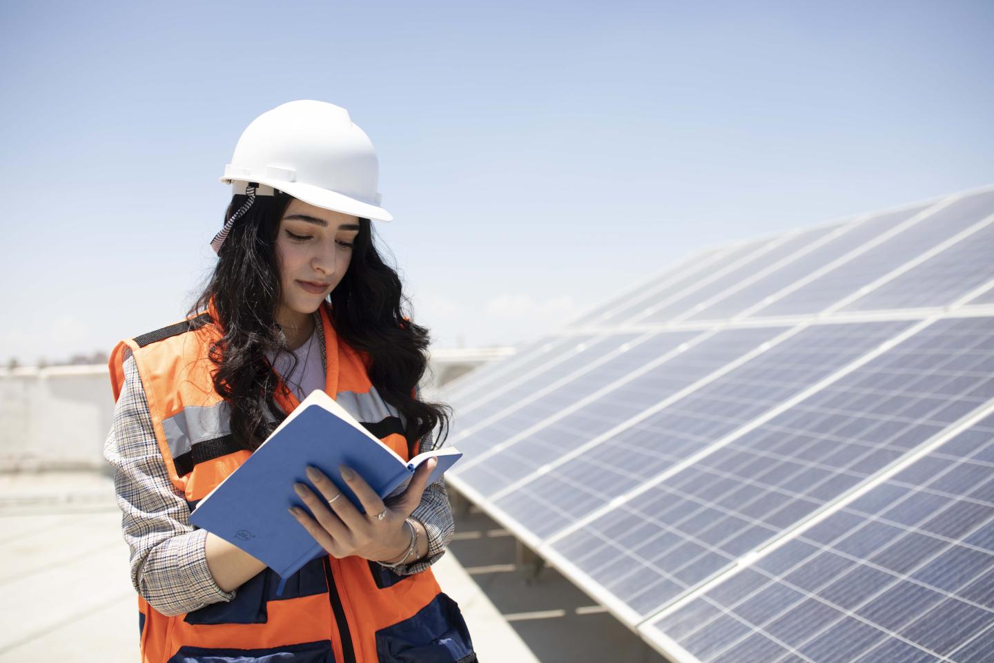 Eine Studentin mit Schutzhelm und Weste steht bei Solaranlagen und notiert etwas in einem Heft.
