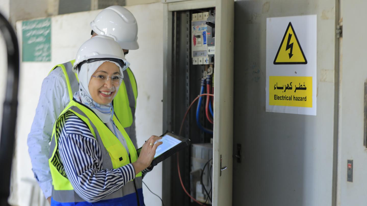 A woman wearing protecting clothing smiles while using a tablet near an electrical panel with a sign indicating an electrical hazard.