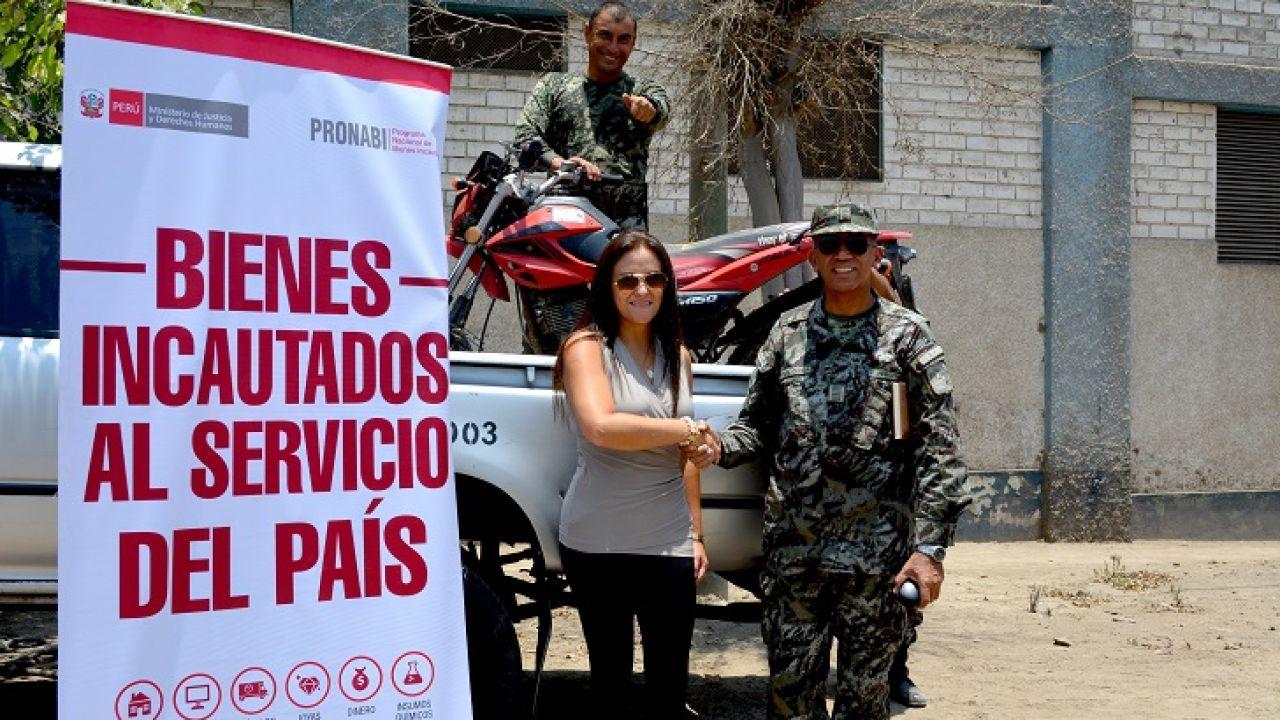 Three people stand next to a seized motorbike that has been loaded onto a truck. There is a sign in the foreground that says ‘Bienes incautados al servicio del país’.