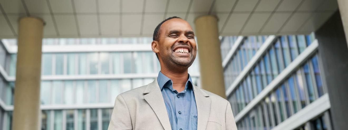 A smiling man in a light-coloured jacket and blue shirt stands in front of a modern office building with a glass façade.