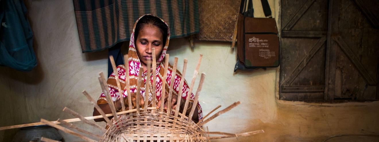 Eine Frau in traditioneller Kleidung sitzt in einem einfachen Innenraum und flechtet konzentriert einen grossen Korb aus Bambus oder ähnlichem Naturmaterial.       ChatGPT fragen
