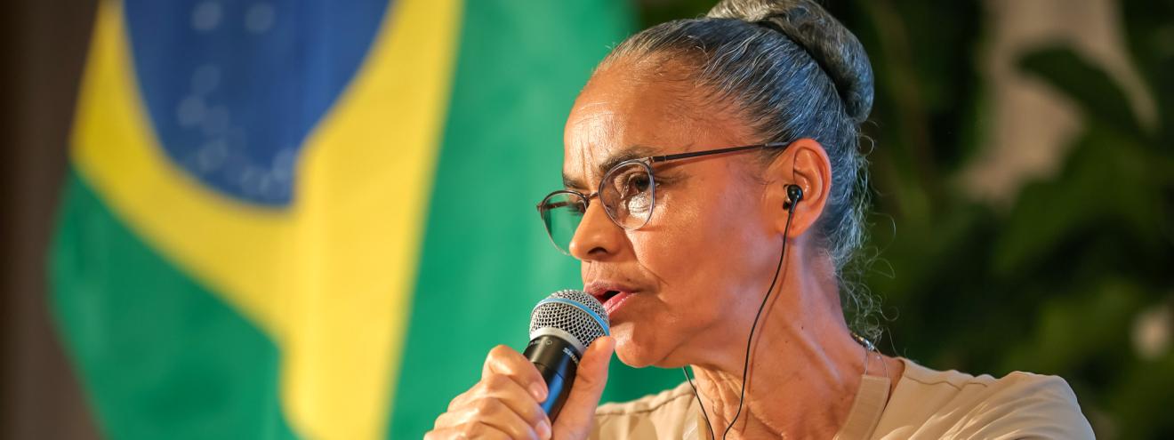 A woman with glasses speaks into a microphone, with the Brazilian flag in the background.