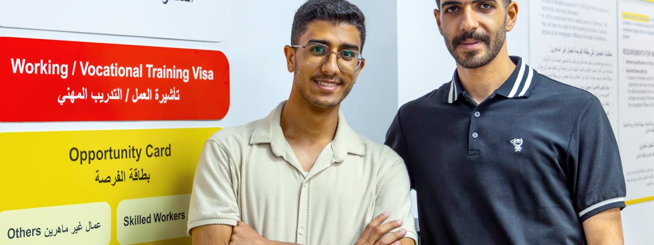 Zwei junge Männer stehen nebeneinander vor einem Informationsplakat zu Visa- und Arbeitsmöglichkeiten in Jordanien.