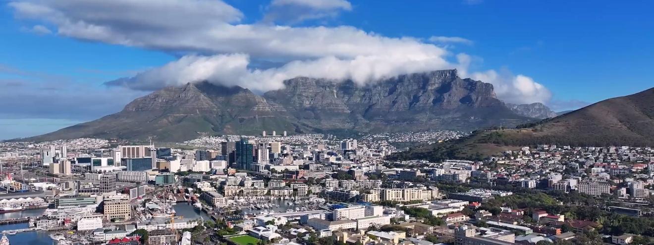Panoramaaufnahme von Kapstadt mit dem Tafelberg im Hintergrund, teilweise von Wolken bedeckt, und dem Stadtzentrum sowie dem Hafen im Vordergrund.