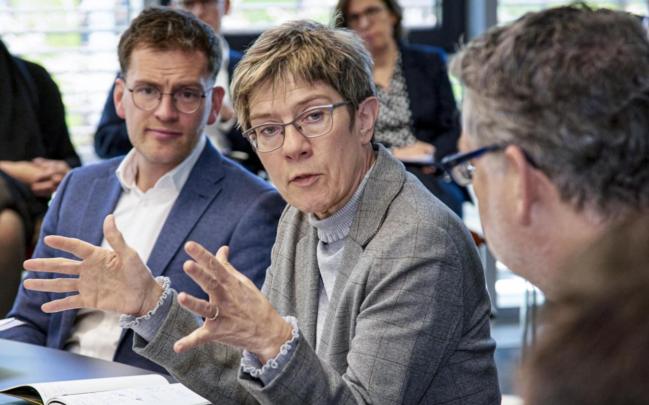 Discussion in a conference room with a woman in a grey blazer speaking animatedly, surrounded by several people in business attire listening attentively.