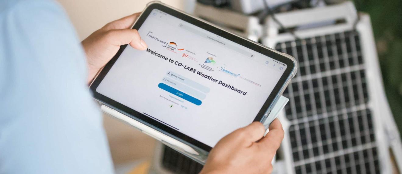 A person holds a tablet with the weather dashboard login page open in front of a solar-powered device.