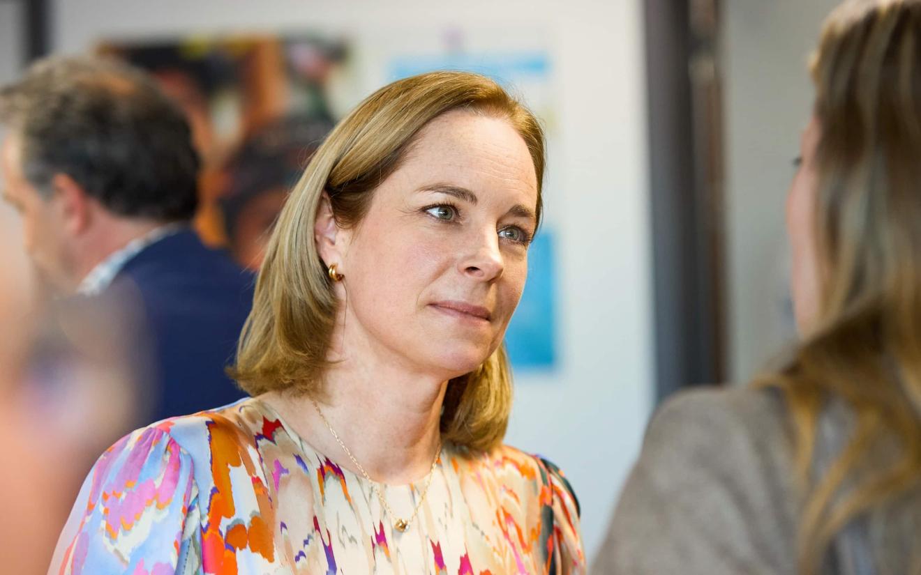 A woman with shoulder-length blonde hair and a colourful blouse talking to another person, with blurred people and pictures on the wall in the background.